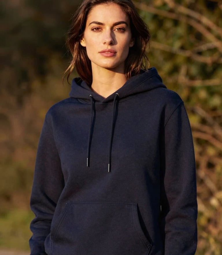 Medium-close portrait of a woman standing confidently in a structured deep navy hoodie, warm golden-hour side light, soft natural foliage background