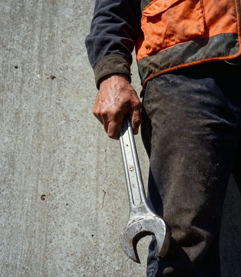 Low-angle heroic close-up of a worker in high-vis gear holding a heavy iron wrench, weathered hands, harsh daylight, concrete background.