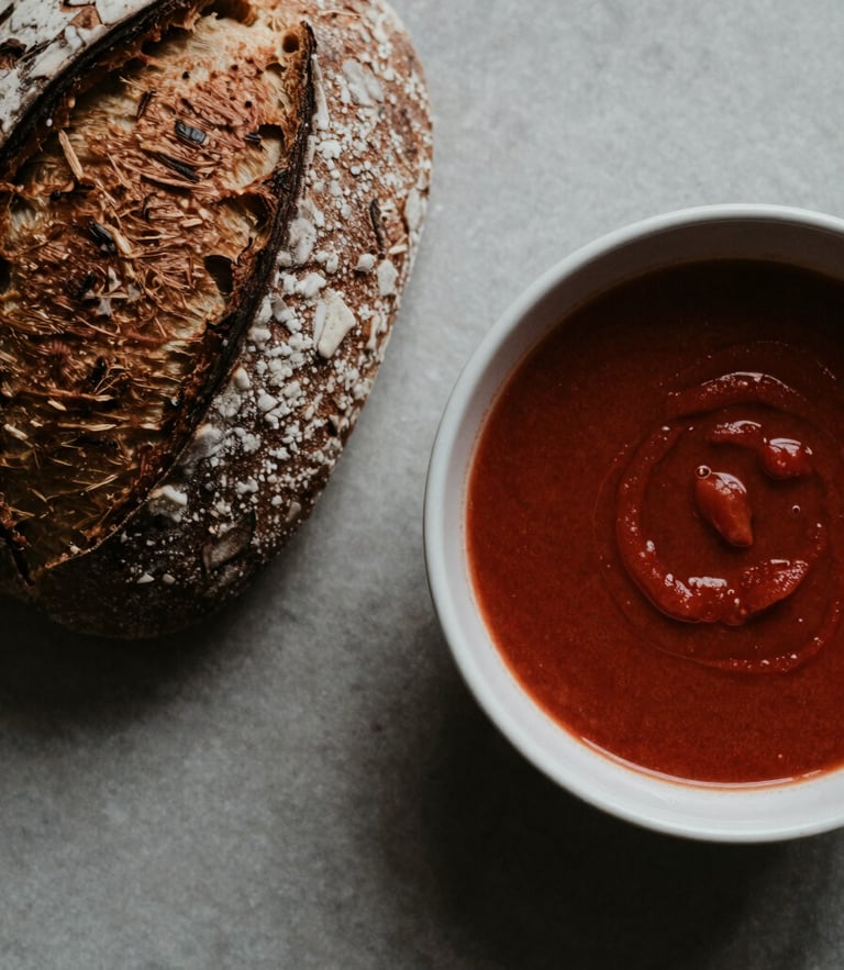 Overhead flat-lay of a rustic sourdough bread next to a ceramic bowl of dark red tomato soup, soft north-facing daylight, moody shadows, editorial food styling