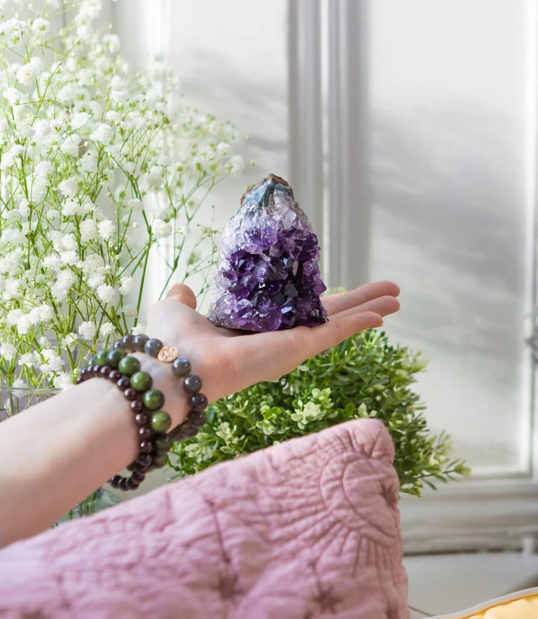 A photo of a hand holding a purple Amethyst cluster crystal with some white flowers