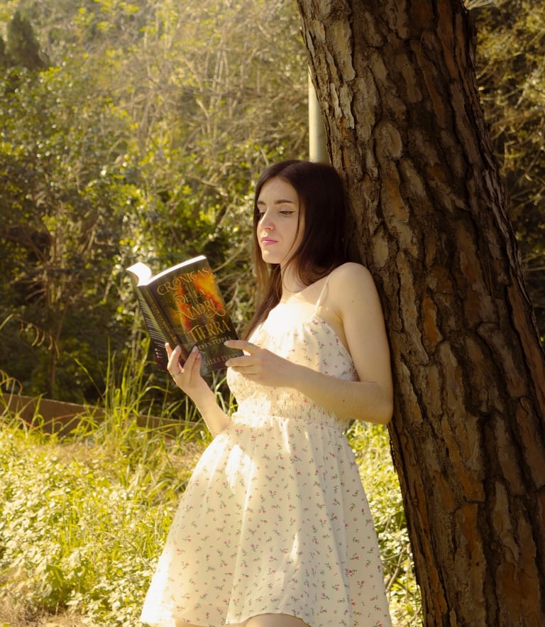 A young woman in a floral summer dress leans against a pine tree reading a fantasy book in a sunlit forest.