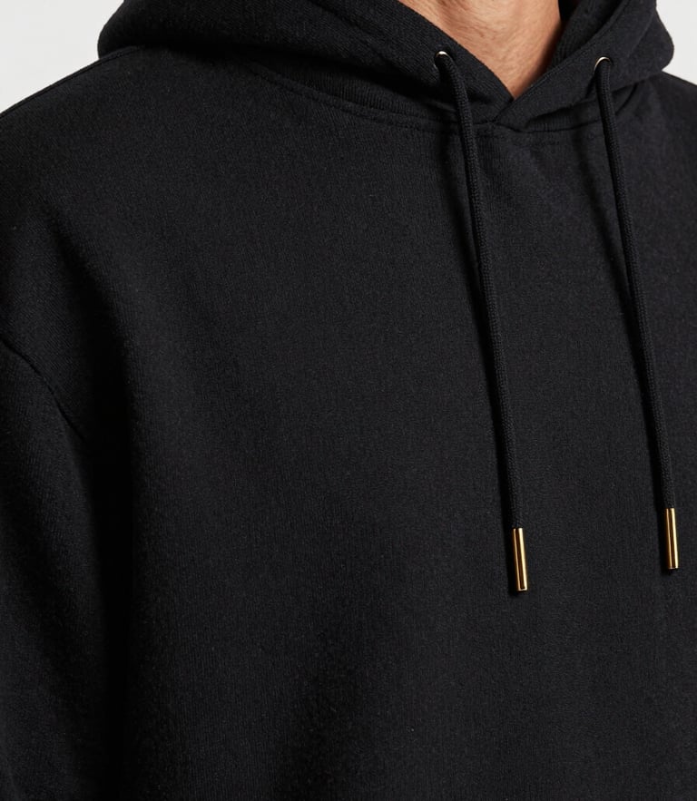 Close-up macro shot of a heavy-drape black cotton hoodie on a model, soft natural shadows, highlighting the thick textile grain and precise stitching, desaturated tones, 35mm lens.