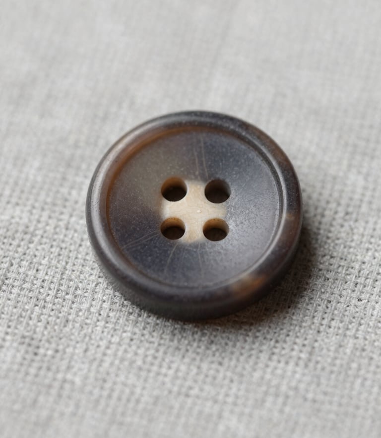 Macro photograph of a single exquisite dark horn button on a light gray linen fabric, showing its unique natural grain and precise 20mm caliber, soft diffused studio lighting.