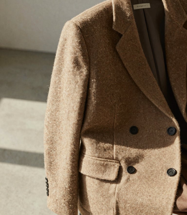 A close-up of a warm oatmeal wool blazer draped over a chair in a sunlit, minimalist concrete studio, soft shadows.