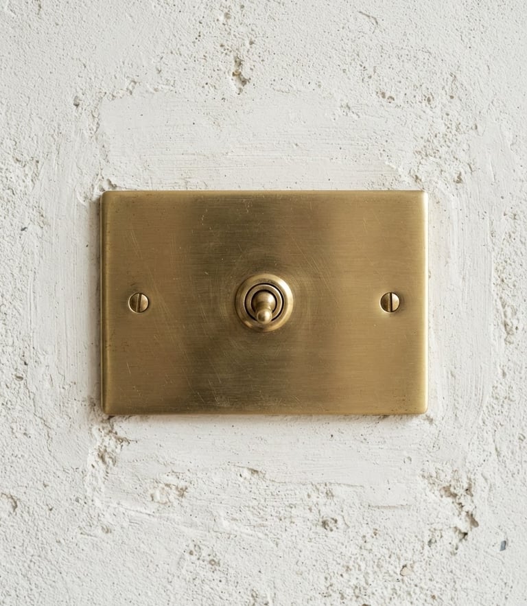 Bright architectural photography of a premium brushed brass light switch mounted on a textured off-white plaster wall. Soft natural daylight casting gentle shadows, sharp macro detail showing the metal grain, 35mm lens, straight-on architectural angle.