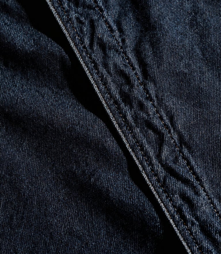 Extreme close-up of heavy raw selvedge denim seams, dark indigo fabric texture, high-contrast directional studio light, deep shadows