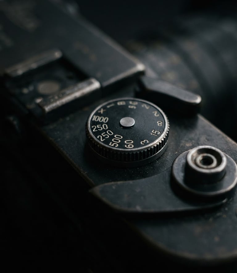 Extreme close-up of a vintage rangefinder camera top plate, focus on the shutter speed dial, low-key side lighting on deep charcoal metal, shallow depth of field