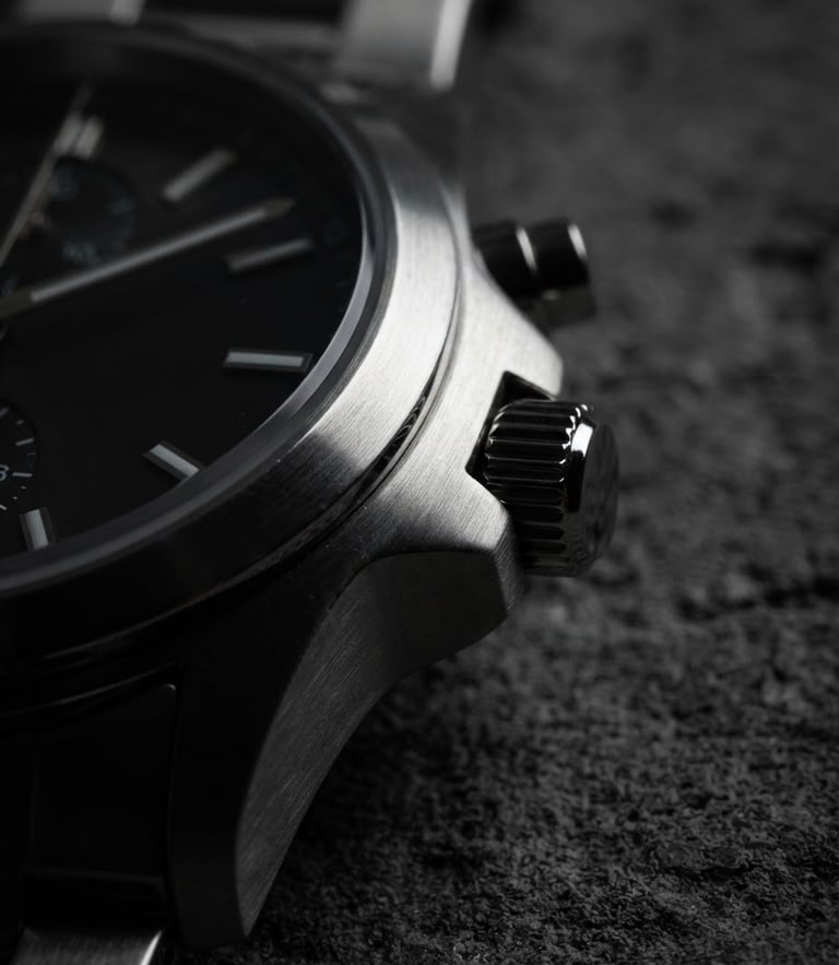 Sharp macro product photography of a brushed steel watch case catching directional light at three o'clock, deep shadows, architectural concrete background
