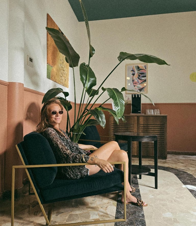 A stylish woman in sunglasses sits in a velvet armchair next to a large bird of paradise plant in a modern cafe.