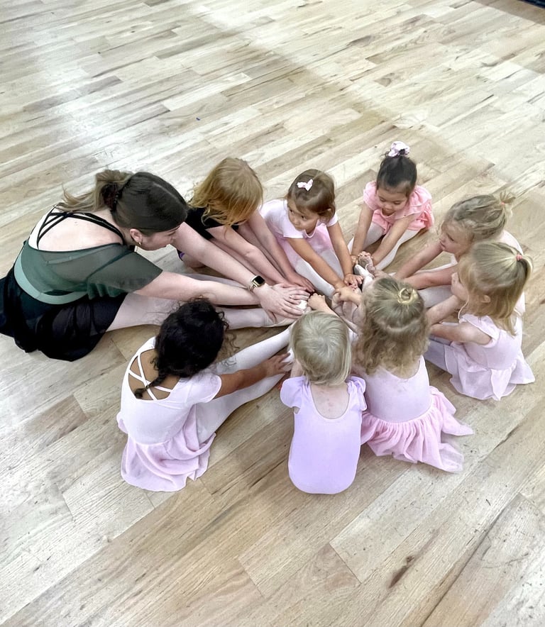 A ballet teacher and young students in pink leotards sit in a circle on a wood studio floor.