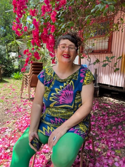 A person with curly brown hair and light skin sits under a tree smiling.