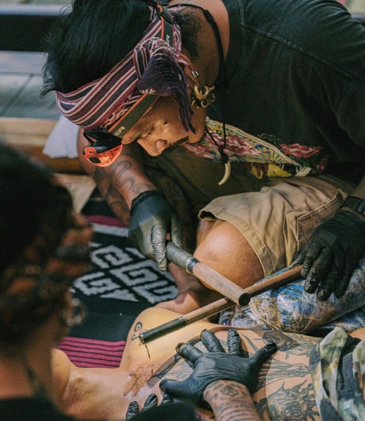 Professional tattoo artist performing a traditional hand-tapped tribal tattoo technique using wooden tools.