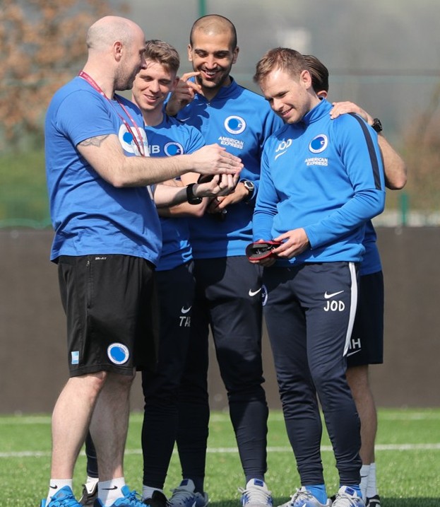 Coach in blue training kit standing with a small group of football staff or players on a pitch, demo