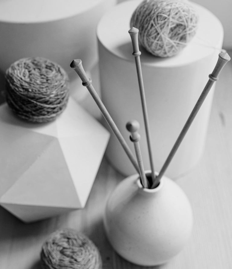 Monochrome still life with textured yarn balls and knitting needles in a vase, alongside white geometric elements..