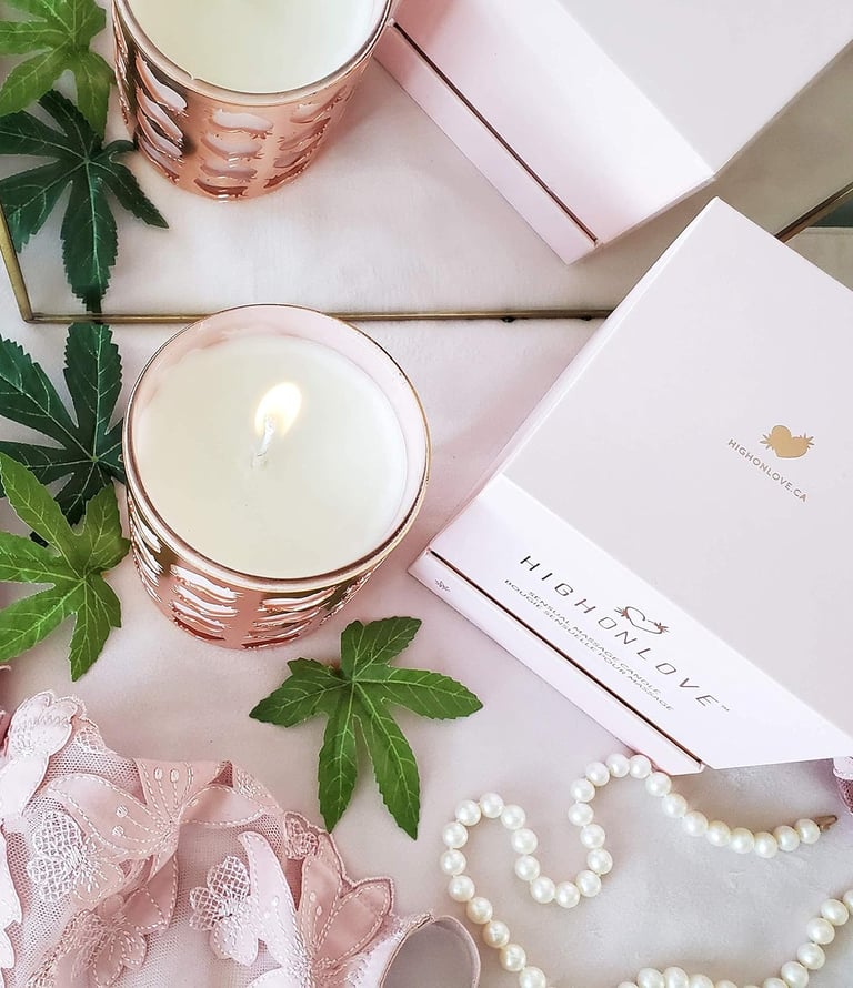 The photo shows a HighOnLove product  - a massage candle - on a background with some pearls, hemp leafs, and the packaging.