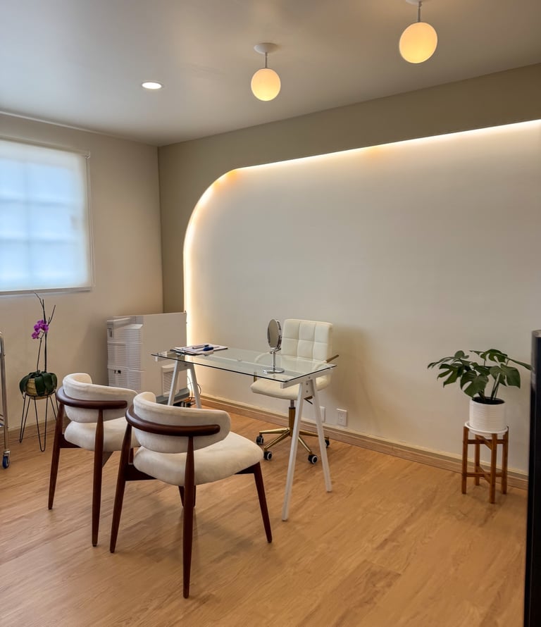 Modern medical spa consultation room with white chairs, glass desk, and warm backlit wall lighting.