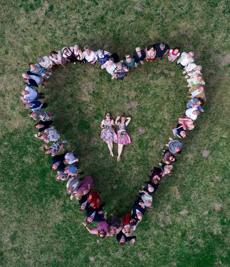 Hochzeitsgesellschaft formt Herz auf Wiese um liegendes Brautpaar. Drohnenaufnahme von Joel Pingel.