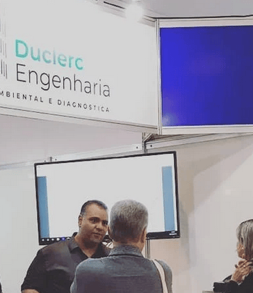 Representante da Duclerc Engenharia atendendo cliente durante feira técnica, apresentando serviços d