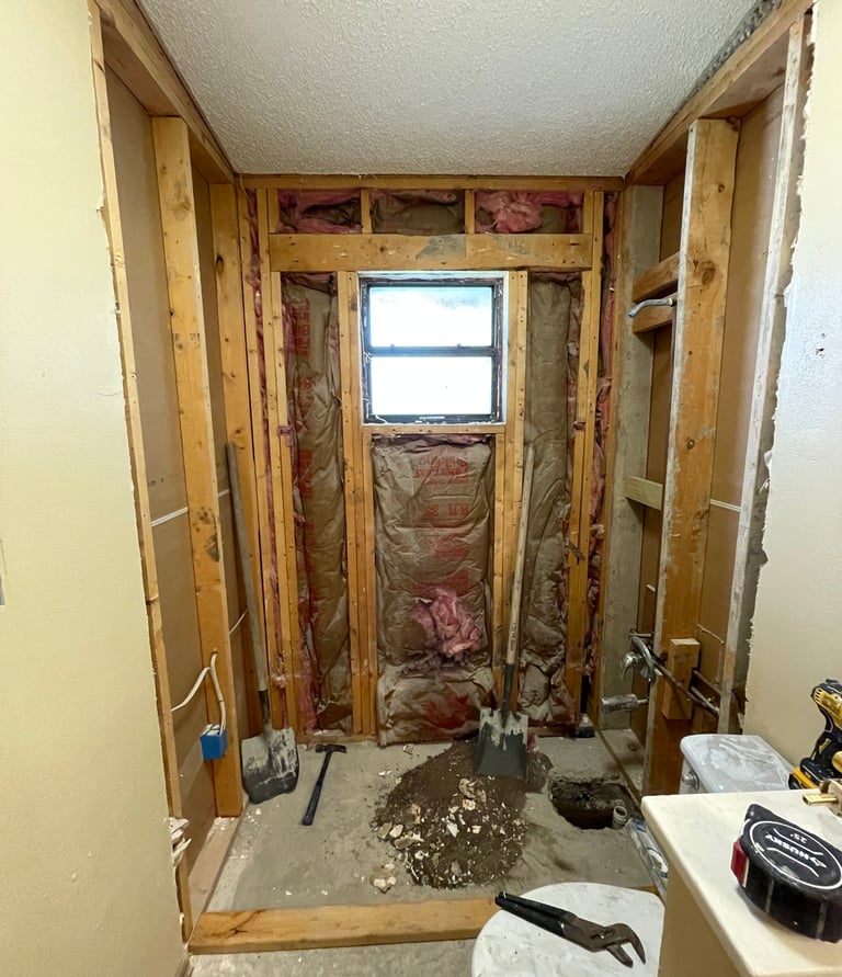 Bathroom renovation project showing exposed wall studs, pink fiberglass insulation, and a small window.