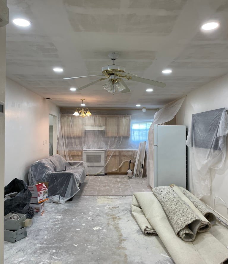Kitchen and living room renovation with plastic-covered furniture, new recessed lighting, and rolled-up carpets.