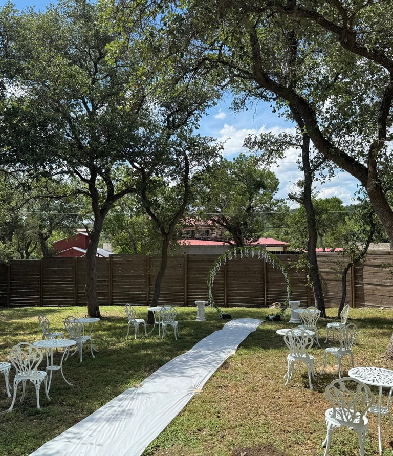 Outdoor wedding ceremony setup with white bistro chairs and an aisle runner under shaded oak trees.