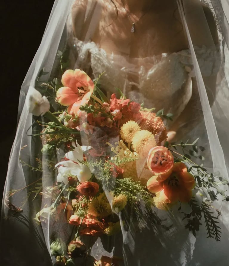 Novia con vestido de encaje bajo velo transparente sosteniendo un ramo en tonos naranja, blanco y verde.