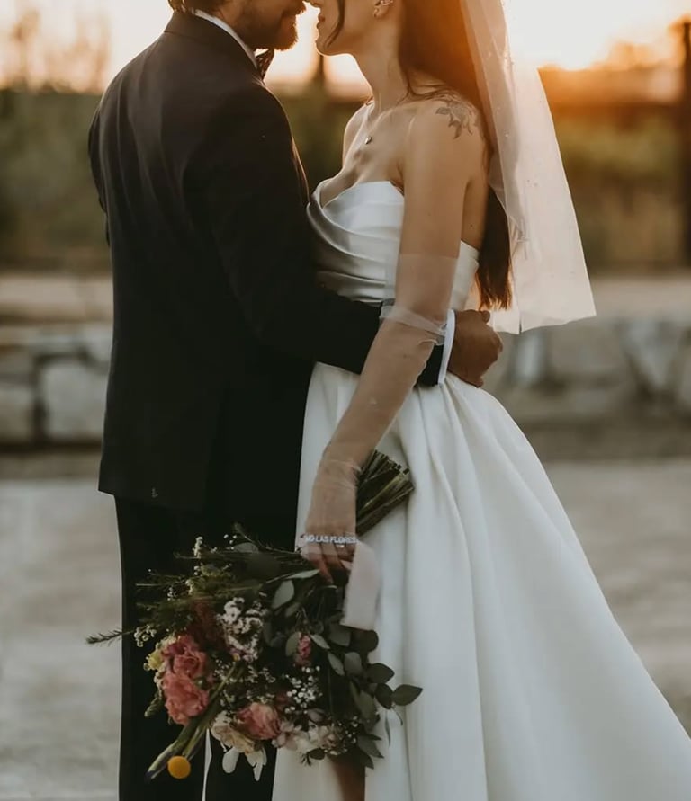 Novios abrazados al atardecer con ramo en tonos rosados.
