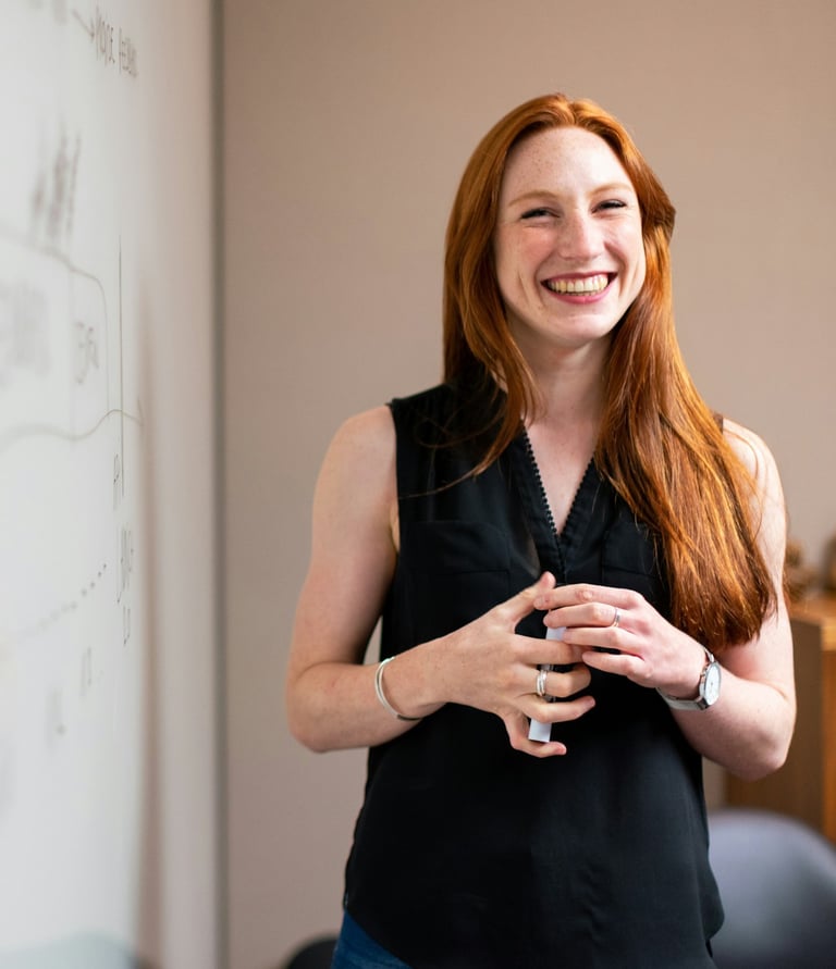 A smiling woman holds a marker and stands next to a white board in the front of a board room.