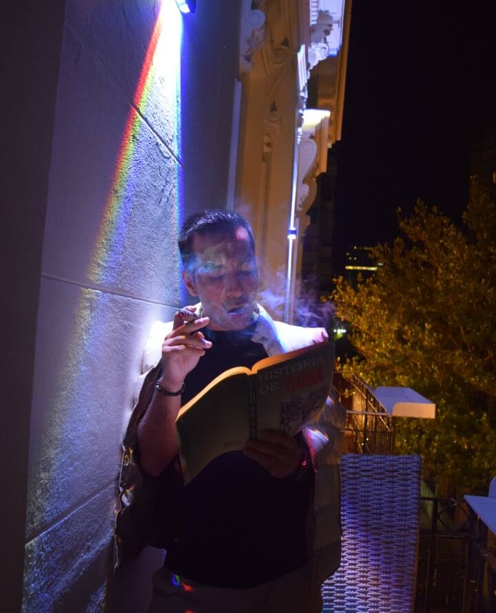 A man smoking a cigar and reading a book on a balcony at night under a rainbow light effect.