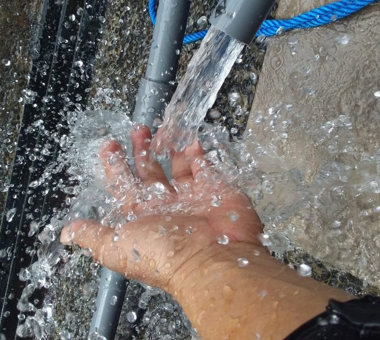 Fresh water splashing onto a person's hand from a grey PVC pipe outdoors.