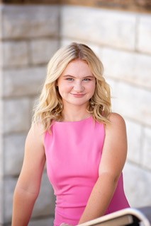 Blonde woman in a pink sleeveless dress posing for a professional portrait against a stone wall.