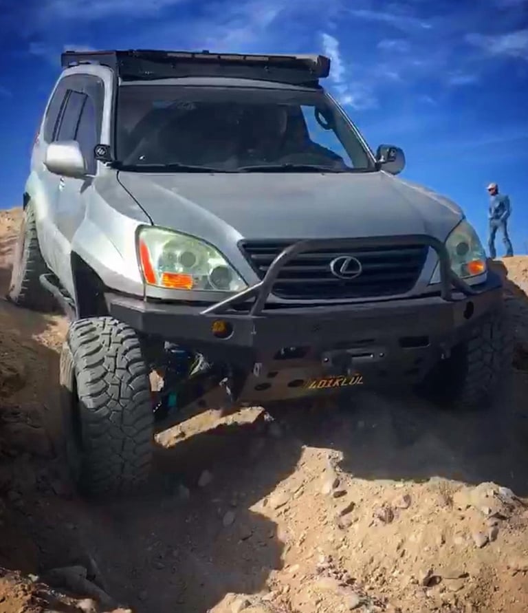 A lifted silver Lexus GX 470 with off-road tires and a steel bumper climbing over rocky desert terrain.