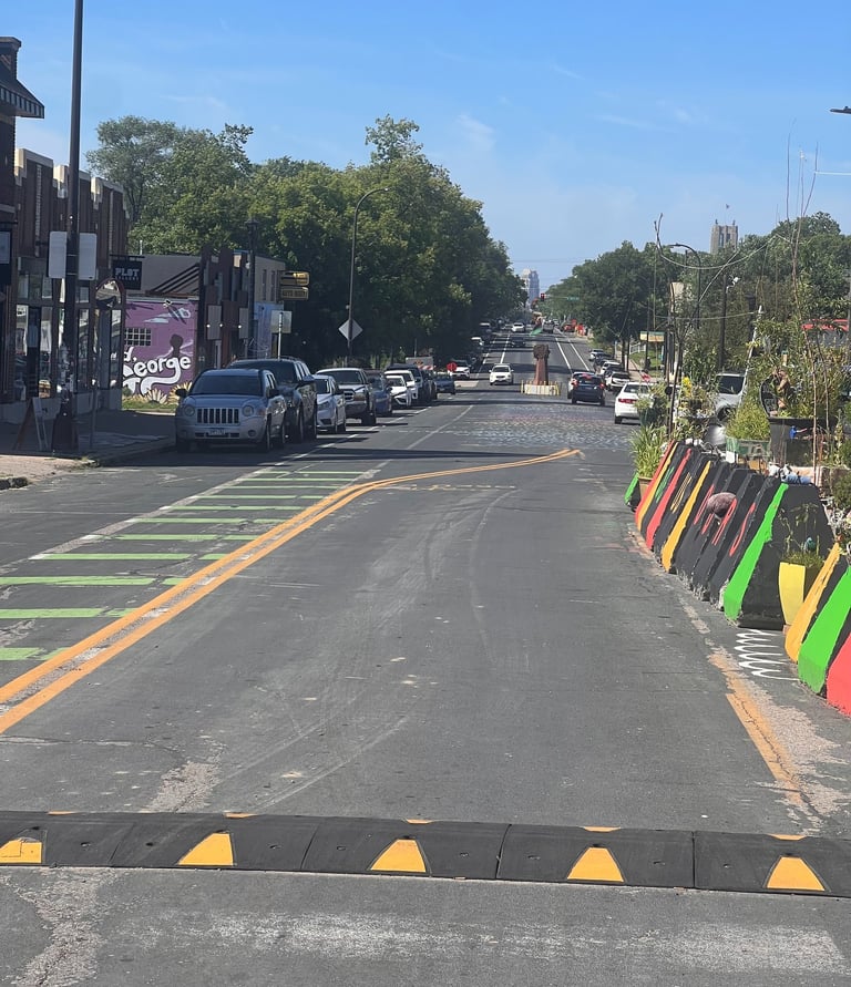 Sunny city street in George Floyd Square with colorful barriers and parked cars under a blue sky.