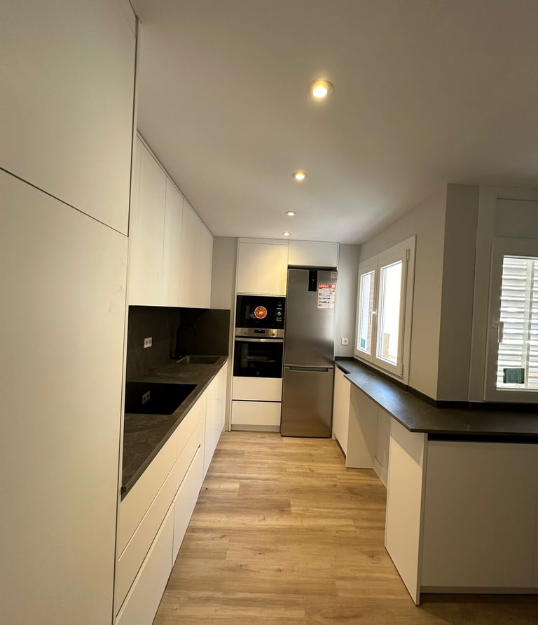 Cocina alargada, muebles blancos, encimeras oscuras, suelo madera.