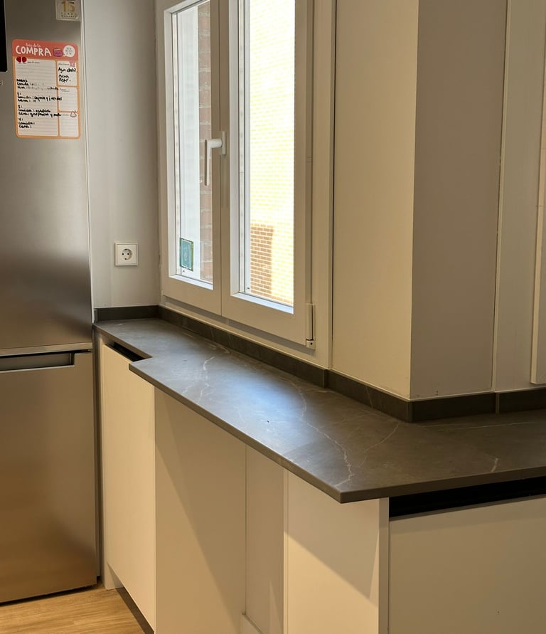 Ventana de cocina, encimera oscura, muebles blancos.