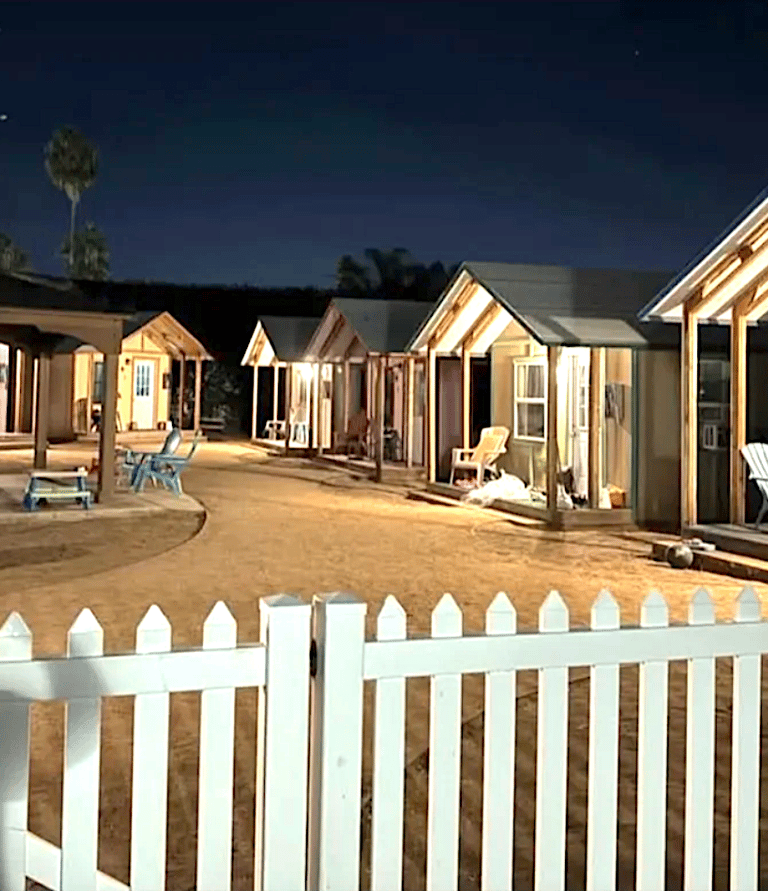 row of hemp town cabins well lit in dusk behind white picket fence