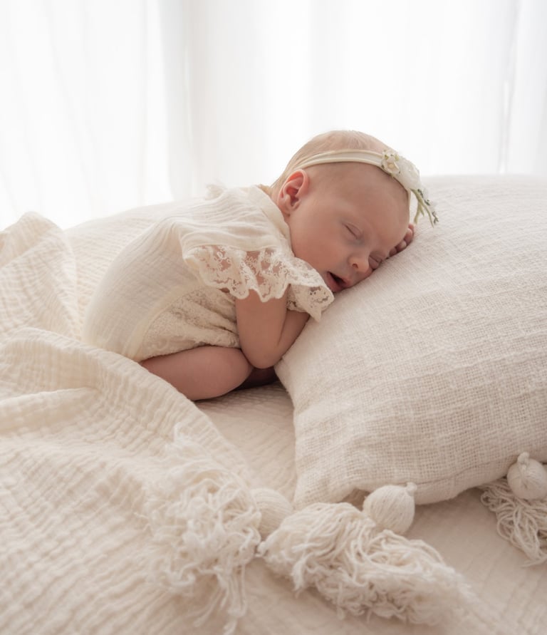 A sleeping newborn girl in a lace romper and flower headband during a professional lifestyle photography session.
