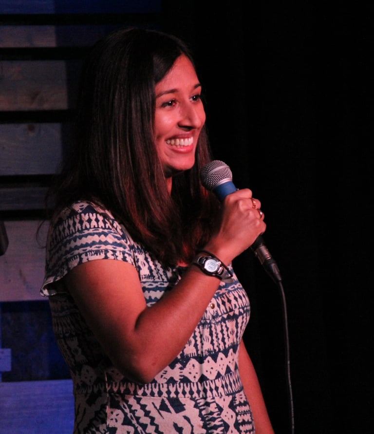 A woman smiles on stage with a microphone, dressed in a patterned dress, in a dimly lit comedy venue.