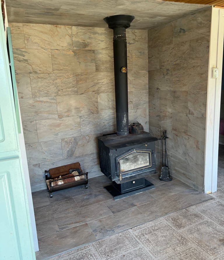 Rustic black wood burning stove on a tile floor with a porcelain wall backdrop and log holder.