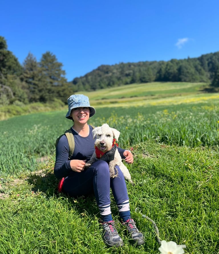 Una mujer sonriente está sentada en un prado verde con su perro blanco mientras hacen senderismo