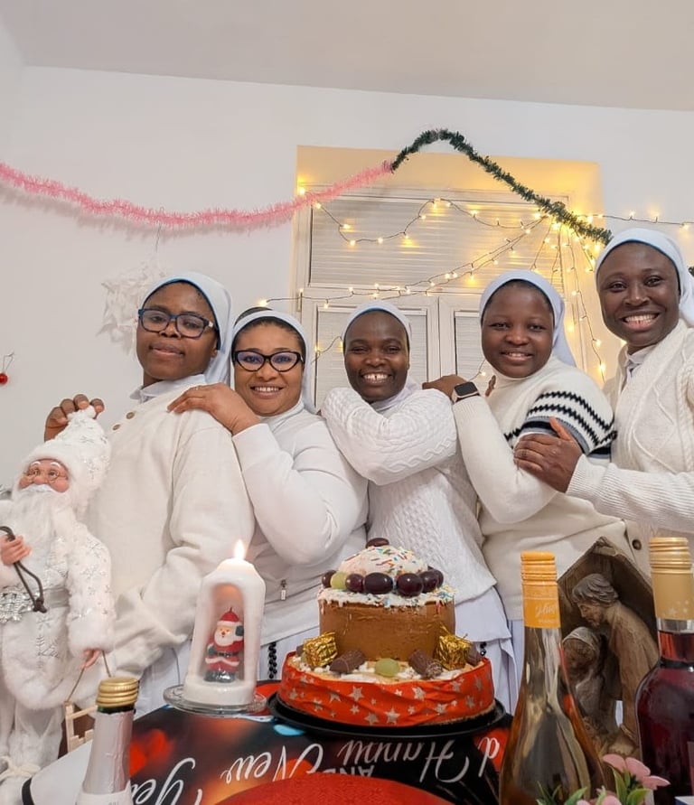 Smiling group of five nuns in white habits celebrating Christmas with a cake, wine, and festive decorations.