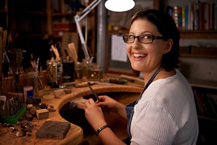 Artesana joyera sonriendo mientras trabaja en una mesa de trabajo de madera con herramientas