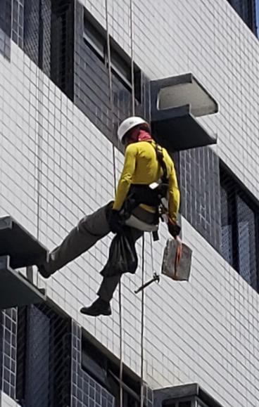 Técnico alpinista realizando teste de percussão e remoção de entulho em fachada predial, durante ins