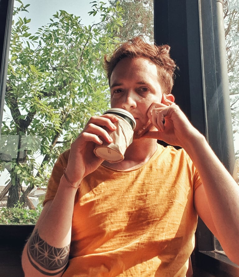 Unicris with a geometric arm tattoo drinking coffee by a sunlit cafe window.