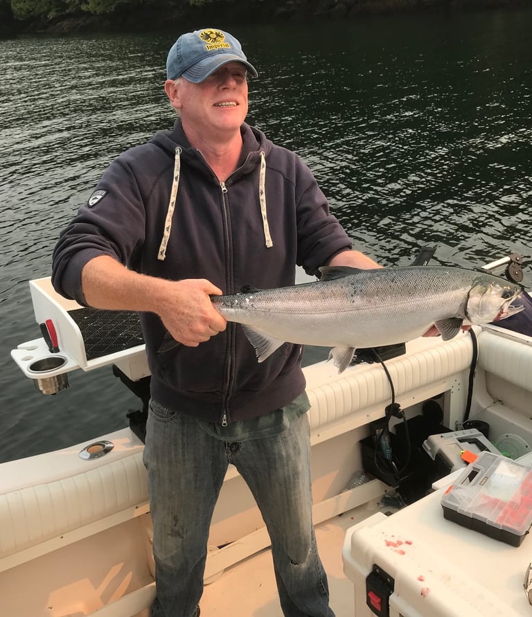 A smiling fisherman holds a freshly caught silver salmon on a boat in calm coastal waters.