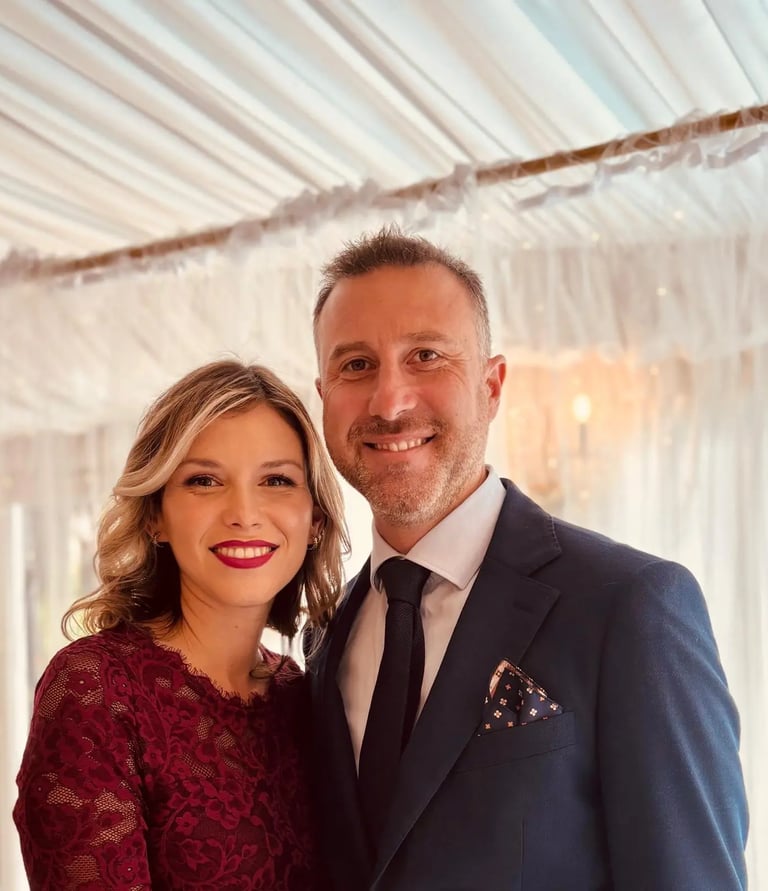 A smiling couple dressed in formal wedding guest attire posing at an elegant indoor event venue.