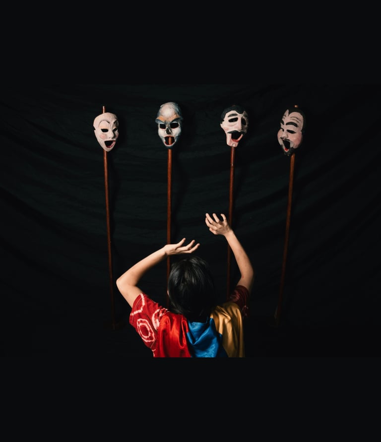 A theatrical performer reaches toward four expressive hand-painted theater masks on wooden poles.