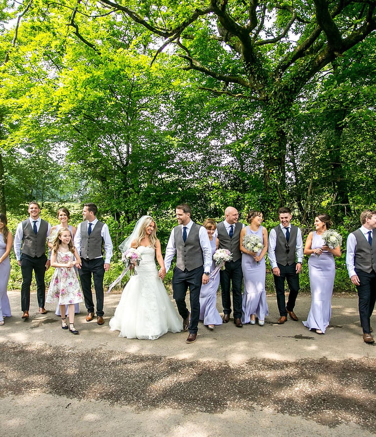 A bride and groom walk with their wedding party down a tree-lined path during a summer woodland wedding.