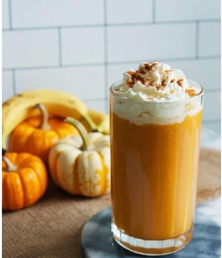 Tall glass of pumpkin smoothie topped with whipped cream and cinnamon, with fresh bananas and small