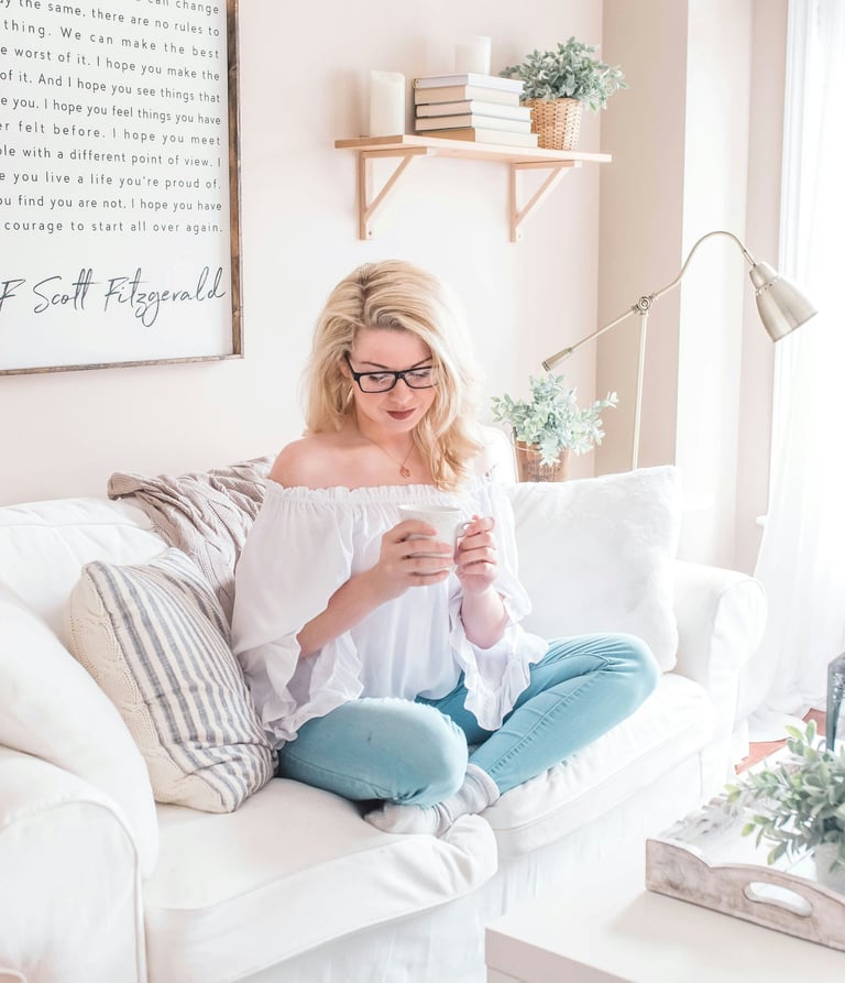 A blonde woman sitting on a white sofa in a bright living room, holding a mug.